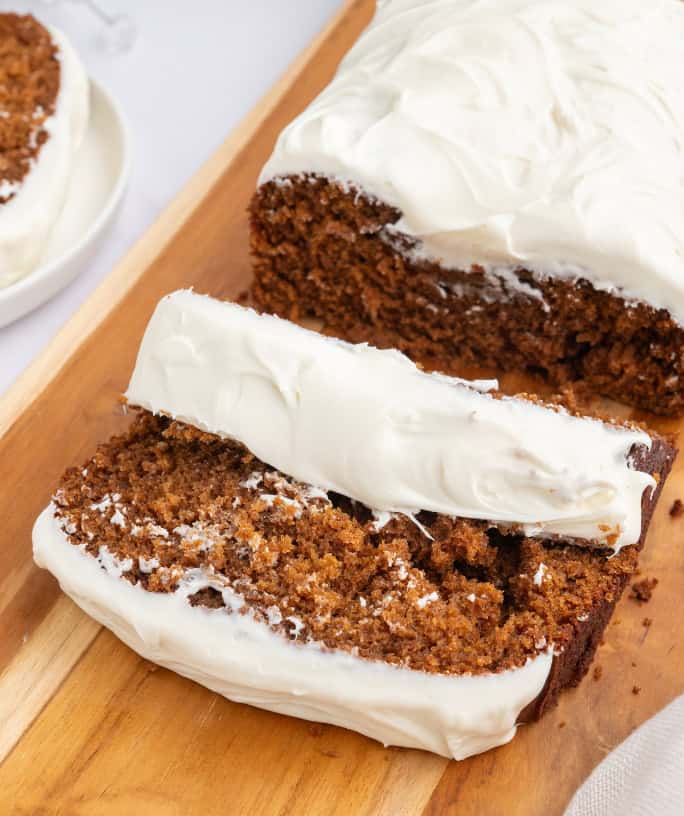 Gingerbread loaf with maple cream cheese frosting, on a cutting board, with two slices cut out of it.