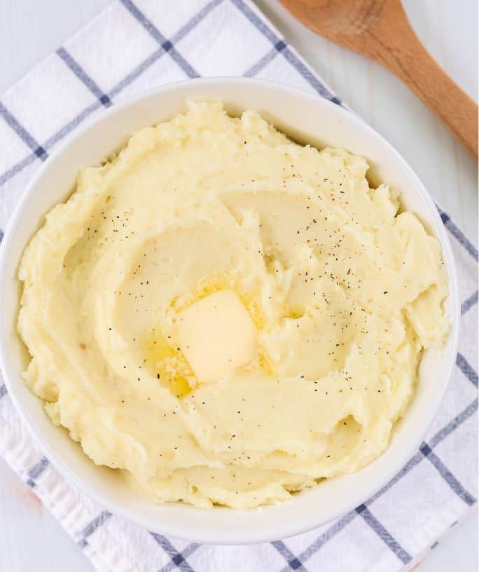 overhead view of a bowl of instant pot mashed potatoes atop a blue and white napkin, with a wooden spoon next to it.