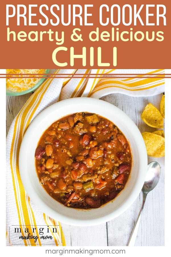 white bowl filled with pressure cooker chili, next to corn chips, a spoon, a white and yellow kitchen towel, and a bowl of shredded cheese