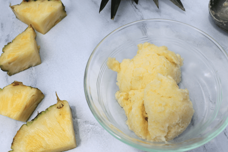 two scoops of pineapple sorbet in a glass bowl, next to slices of pineapple