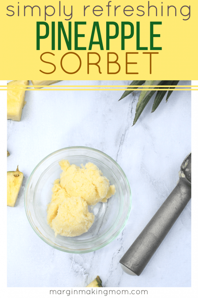 Clear glass bowl with two scoops of homemade pineapple sorbet, next to an ice cream scoop and slices of pineapple