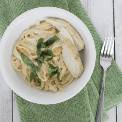 White plate with fettucine alfredo and sliced chicken breast, topped with fresh basil, all resting on a green napkin.