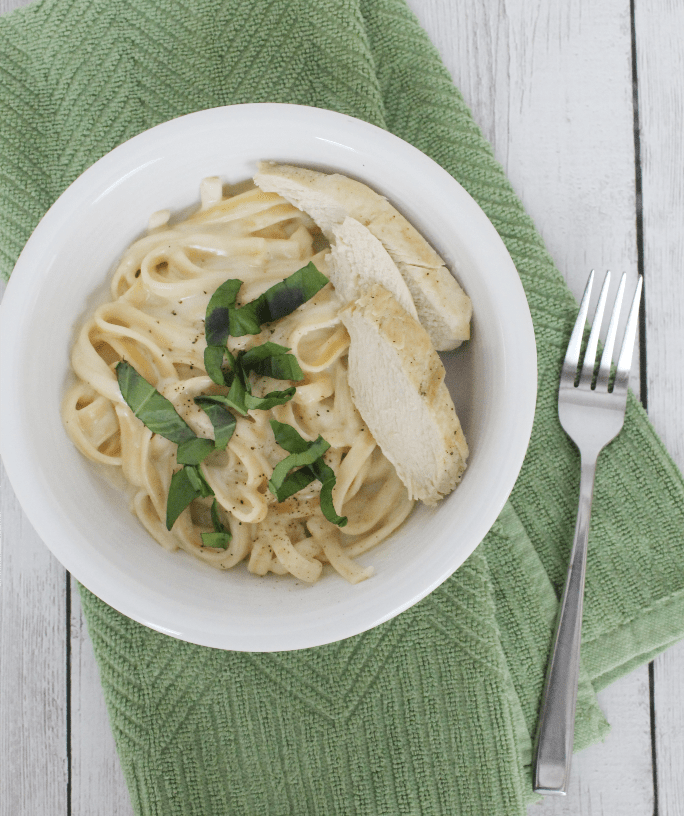 White plate with fettucine alfredo and sliced chicken breast, topped with fresh basil, all resting on a green napkin.