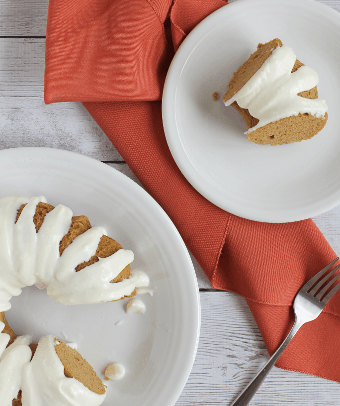 slice of pressure cooker pumpkin spice bundt cake ona white plate, accompanied by the whole cake on another serving plate. Orange cloth napkin under plates.