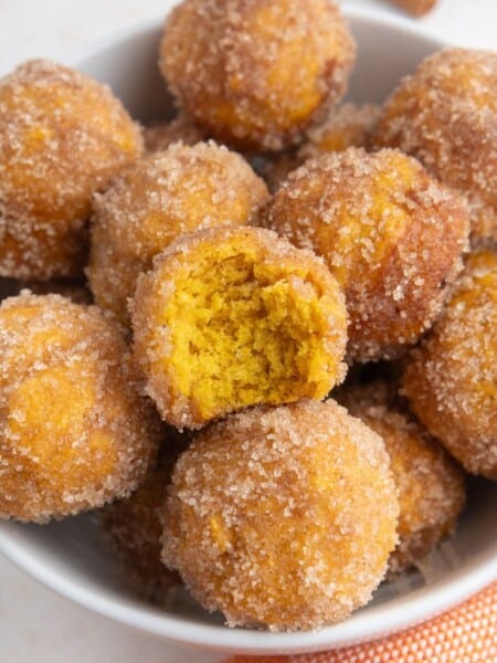 several Bisquick pumpkin donut holes, rolled in cinnamon sugar, served in a white bowl. One has a bite taken out of it, showing its moist pumpkin spice flavored interior.