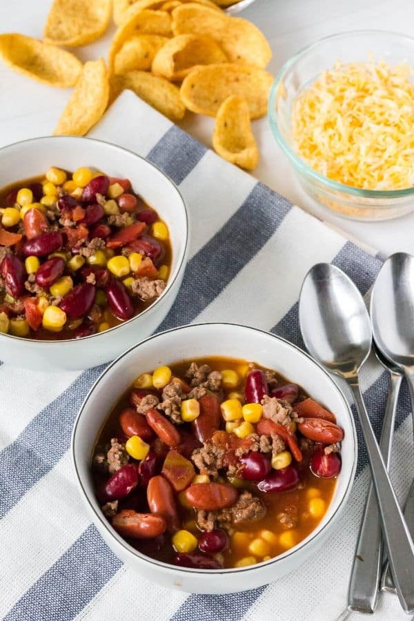 two bowls of Instant Pot taco soup on a blue and white cloth with some spoons, cheese, and frito corn chips