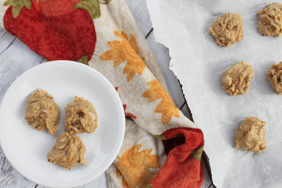 white plate with three pumpkin pecan caramel chip cookies resting on it, set on top of a pumpkin printed hand towel, next to a parchment lined baking sheet of additional cookies