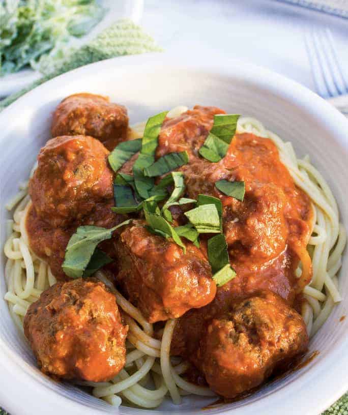 white bowl filled with spaghetti and homemade meatballs in a red sauce, topped with chopped basil