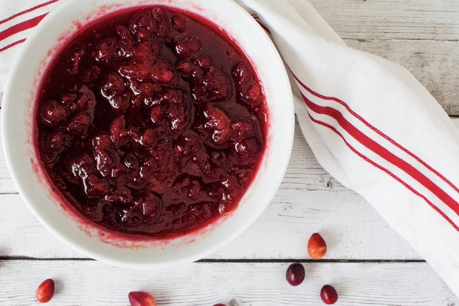 white bowl filled with pressure cooker cranberry sauce, surrounded by scattered fresh cranberries, next to a white and red kitchen towel