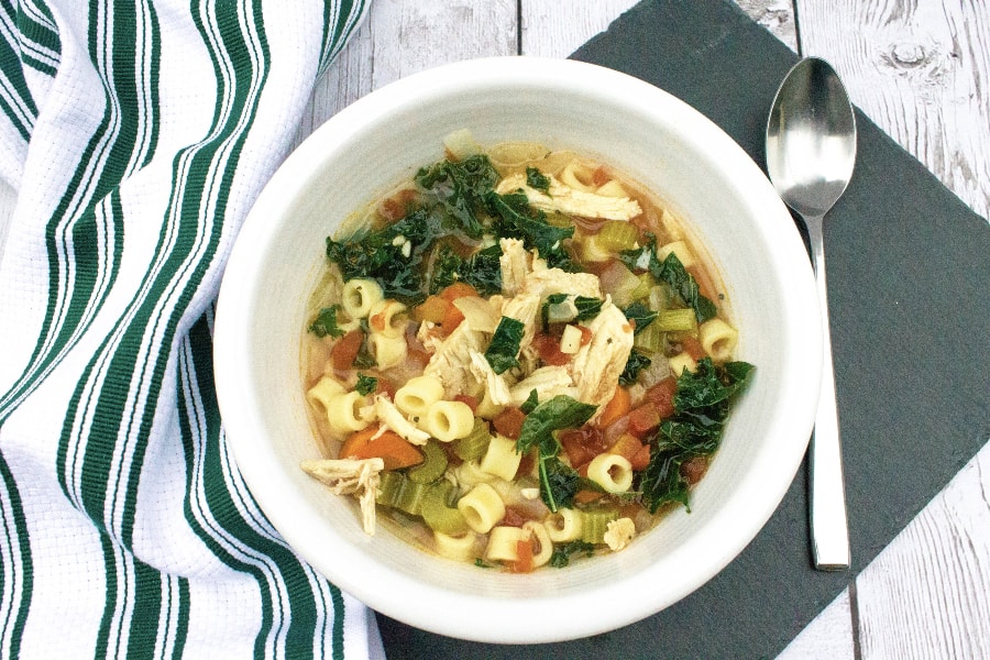 Instant Pot Italian Chicken Soup with vegetables and kale in a white bowl next to a green and white kitchen towel