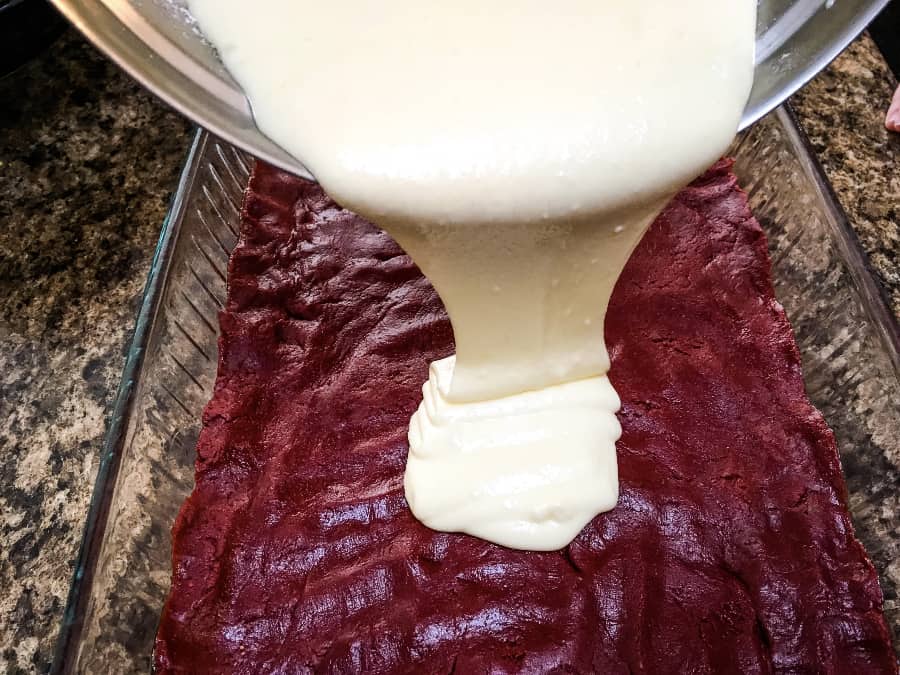 cream cheese filling being poured over the cake mix base to prepare red velvet ooey gooey butter cake dessert bars