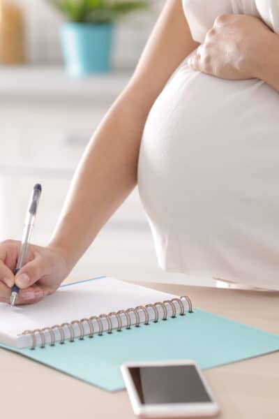 Pregnant woman writing third trimester checklist for maternity in kitchen, closeup