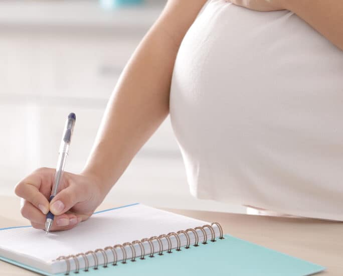 Pregnant woman writing third trimester checklist for maternity in kitchen, closeup