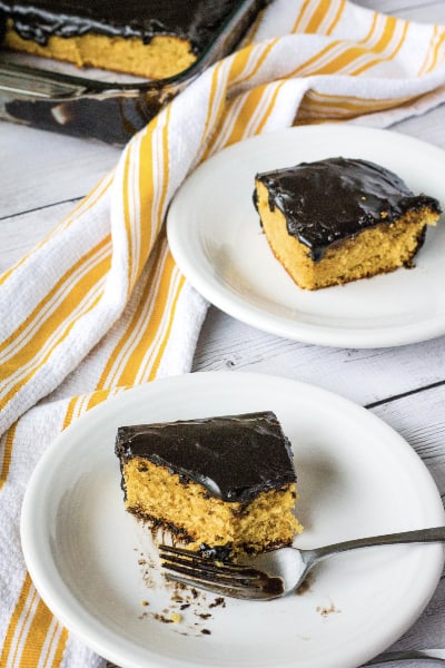 two white plates each with a piece of peanut butter cake with chocolate frosting, next to a yellow and white striped kitchen towel