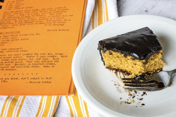 white plate with a piece of peanut butter cake on it, next to a church cookbook with Grandma's peanut butter cake recipe showing