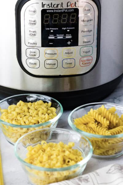 three glass bowls of dried pasta resting in front of an Instant Pot pressure cooker, demonstrating types of pasta that can be cooked in the Instant Pot