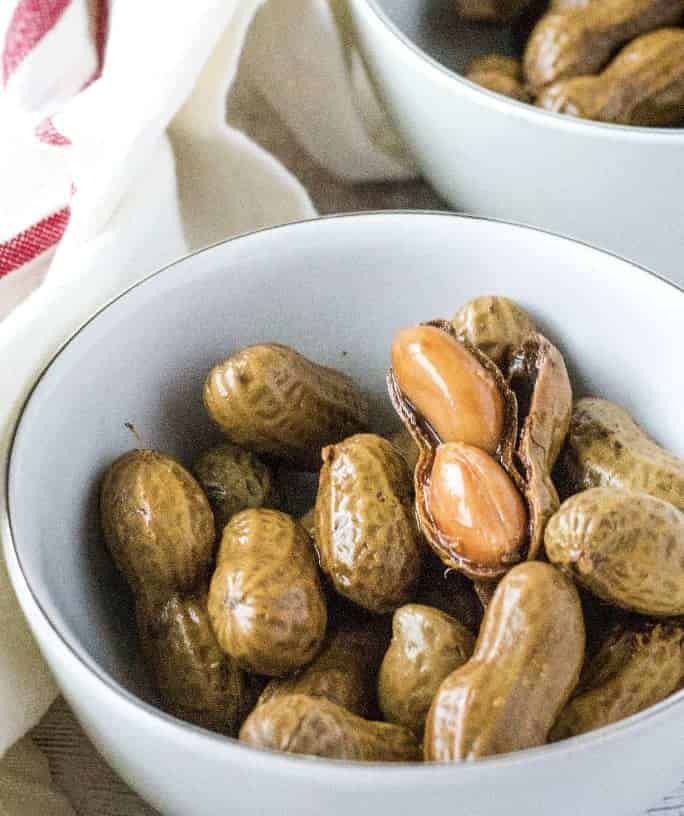 white bowl filled with boiled peanuts in the shell, with one shell opened to reveal the soft peanuts inside