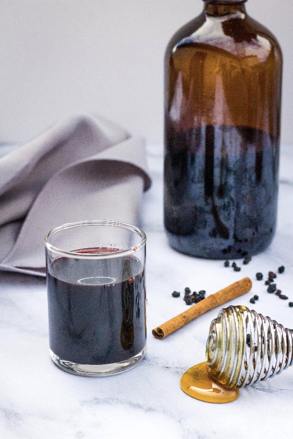 A bottle of elderberry syrup next to a small glass of elderberry syrup, alongside a cinnamon stick and some honey
