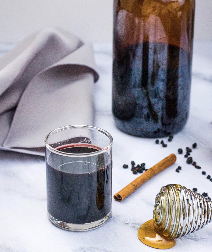 small glass of elderberry syrup next to a larger amber bottle of elderberry syrup