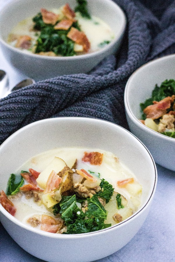 three white bowls filled with healthy zuppa toscana soup, with potatoes, sausage, and kale, topped with bacon crumbles