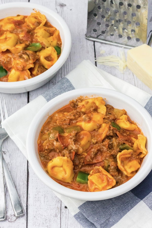 two white bowls of pizza tortellini that was cooked in the pressure cooker, with a cheese grater and block of cheese next to them