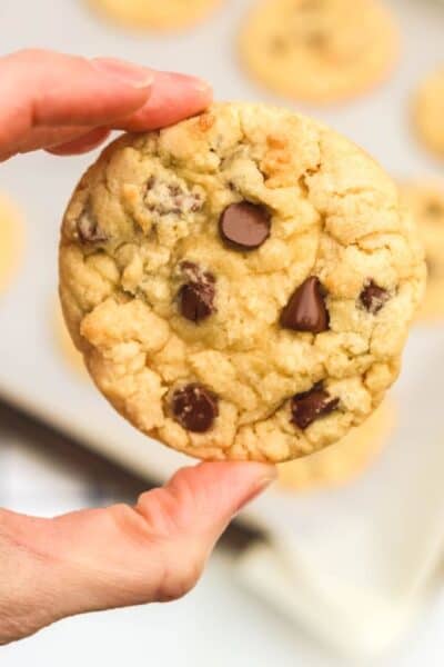 a Bisquick chocolate chip cookie is held by a woman's hand, with the baking sheet of remaining cookies in the background.
