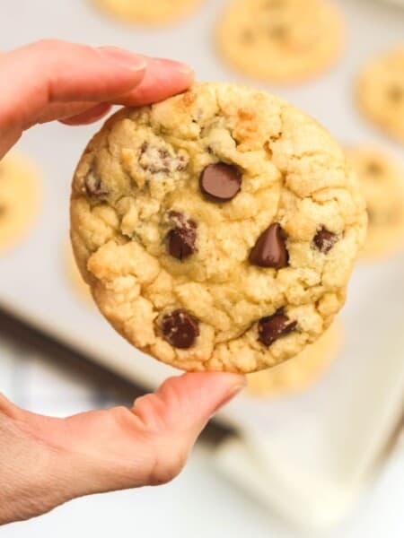 a Bisquick chocolate chip cookie is held by a woman's hand, with the baking sheet of remaining cookies in the background.
