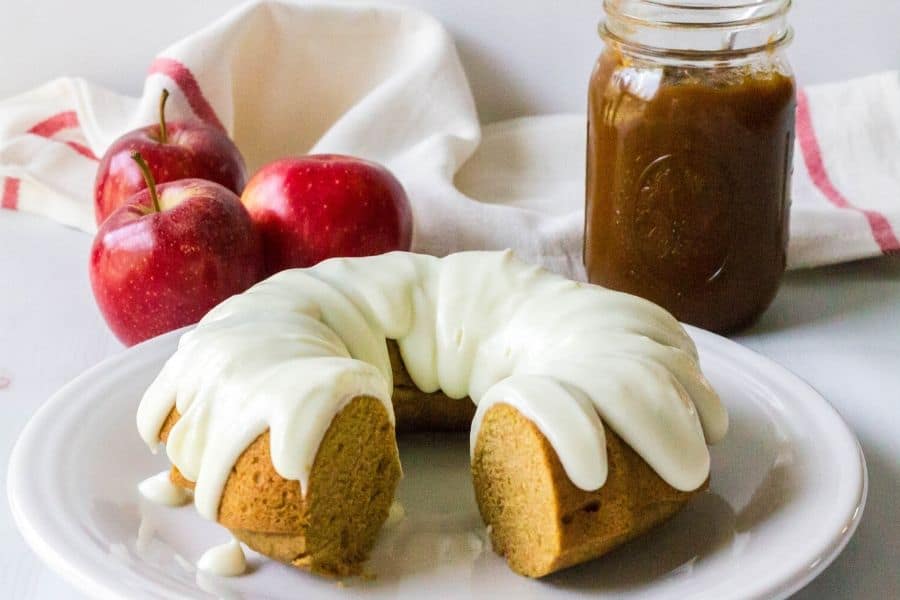 partially cut apple butter bundt cake on a white plate, next to a jar of apple butter and some apples