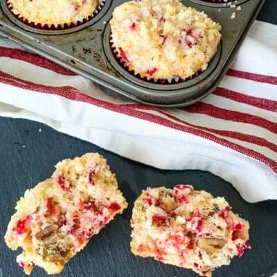 a cranberry orange muffin cut in half, laying on a black slate board next to a muffin pan