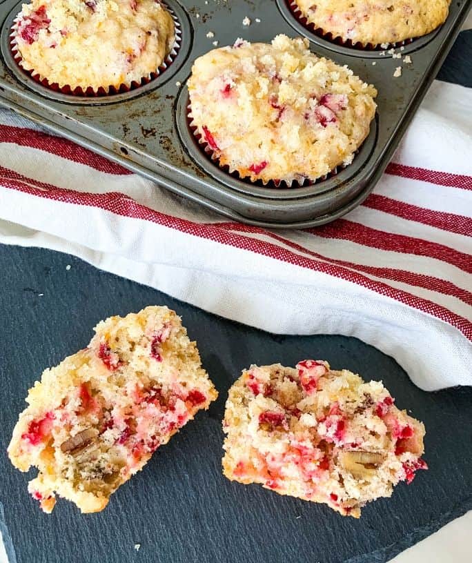 a cranberry orange muffin cut in half, laying on a black slate board next to a muffin pan