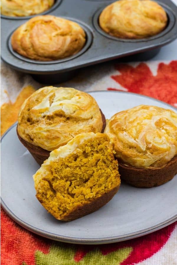 three pumpkin cream cheese muffins on a white plate, with one muffin cut in half to show its soft and moist interior