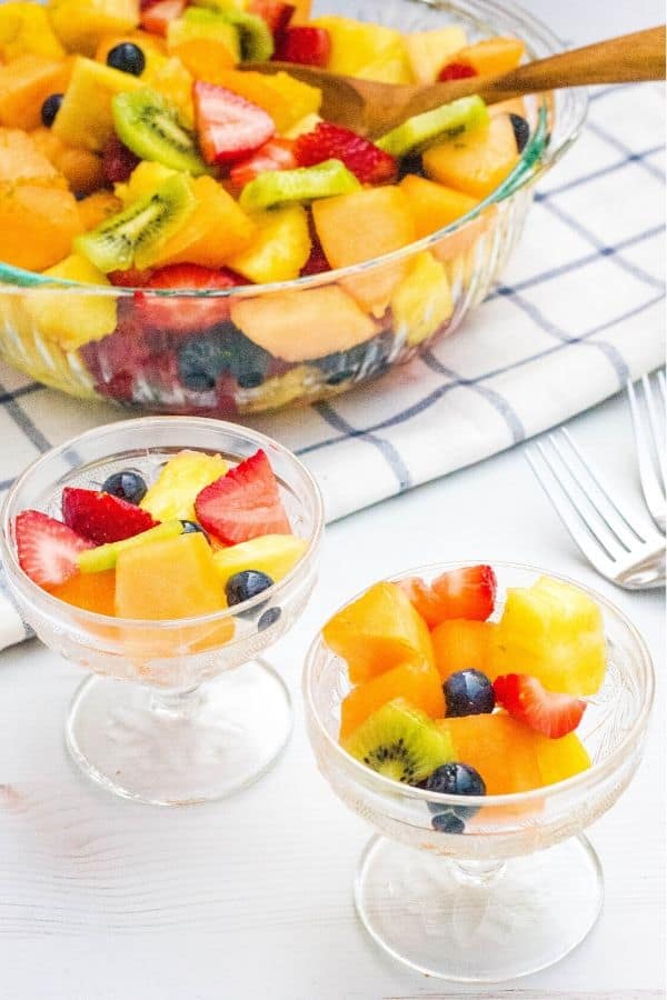 two glass dessert bowls filled with honey lime fruit salad, next to the whole bowl of fruit salad