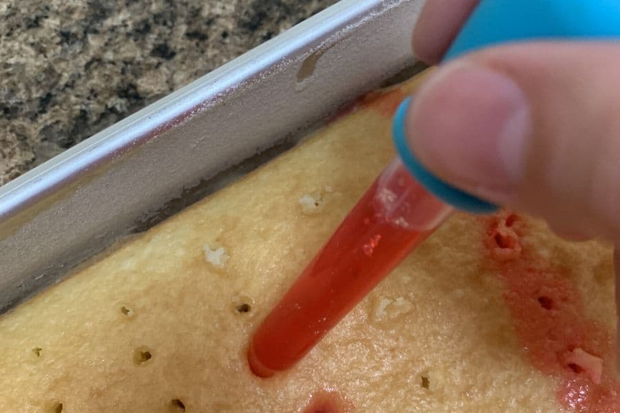 a hand adding red jello to the holes of the poke cake, using a dropper
