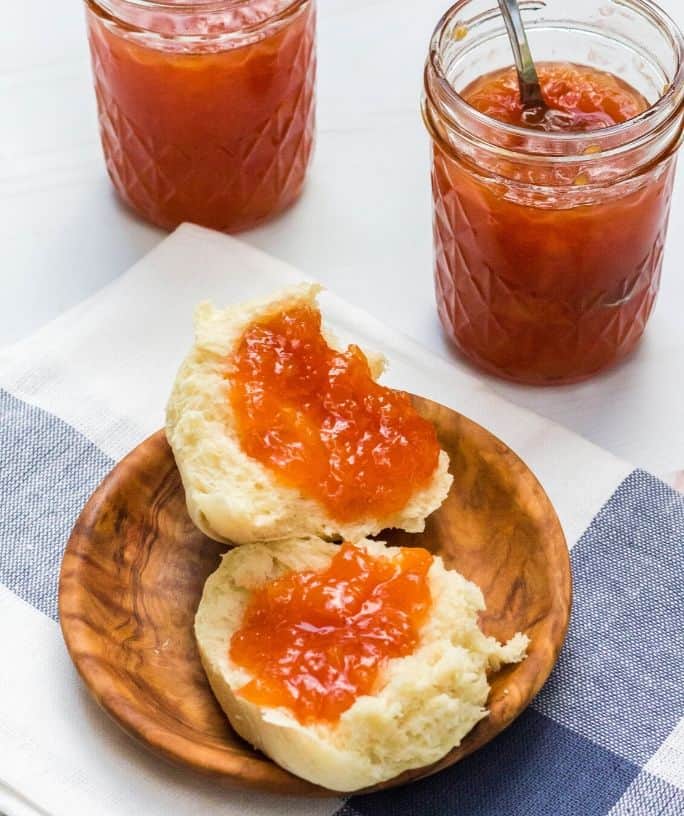 dinner roll with peach jam on it in front of two jars of peach jam