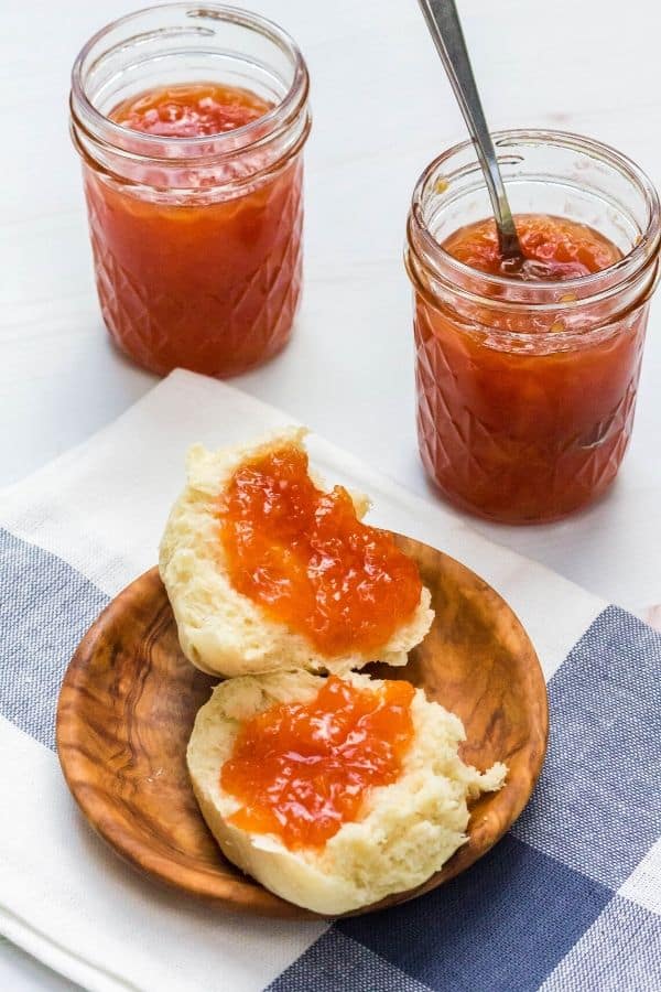 peach jam on a split dinner roll on a wooden plate, in front of two jars of jam