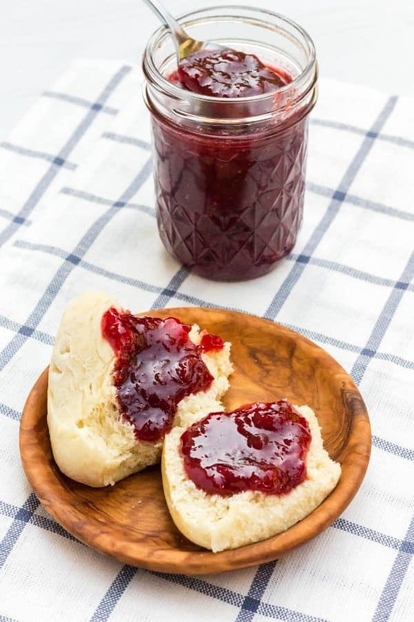 a small wooden plate with a dinner roll on it, with strawberry jam on the dinner roll and the jar of jam in the background