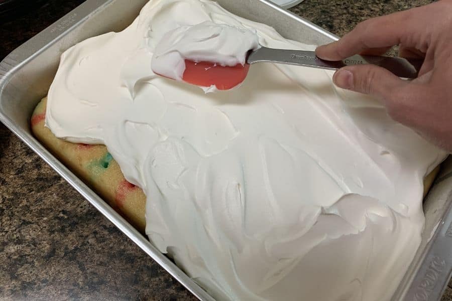 hand holding a spatula and spreading whipped cream onto a red white and blue poke cake