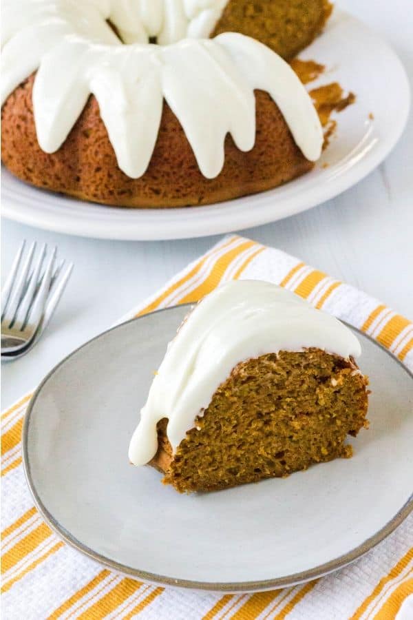 a pumpkin spice bundt cake in the background with a slice of the cake on a plate in the foreground, resting on a yellow and white striped towel