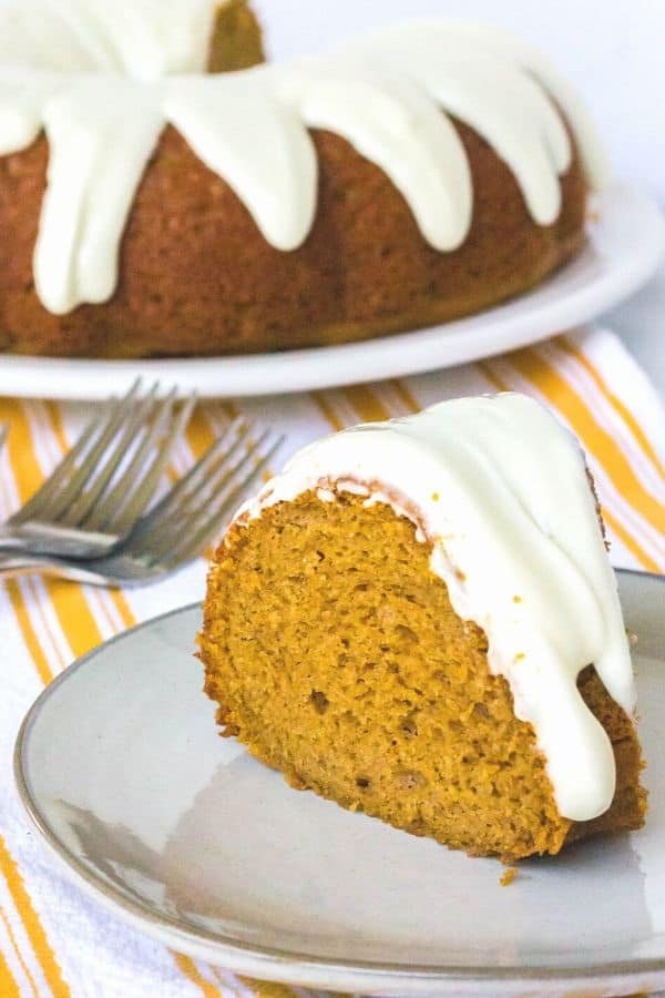 slice of pumpkin bundt cake topped with cream cheese frosting, on an ivory plate in front of the remainder of the cake