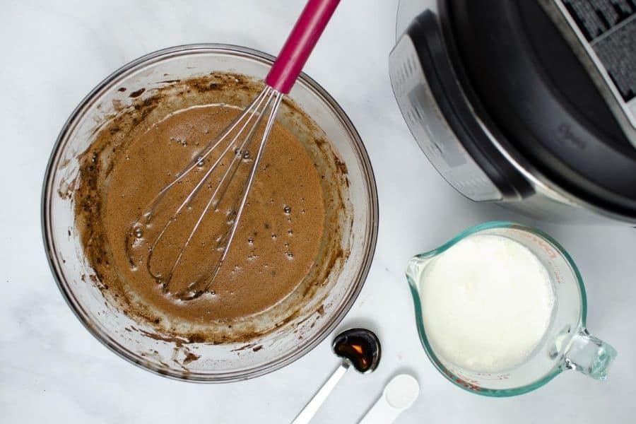 a mixing bowl of cocoa powder, powdered sugar, water, and egg yolks mixed together, next to a measuring cup of cream and a spoon of vanilla extract--all for making pressure cooker pots de creme