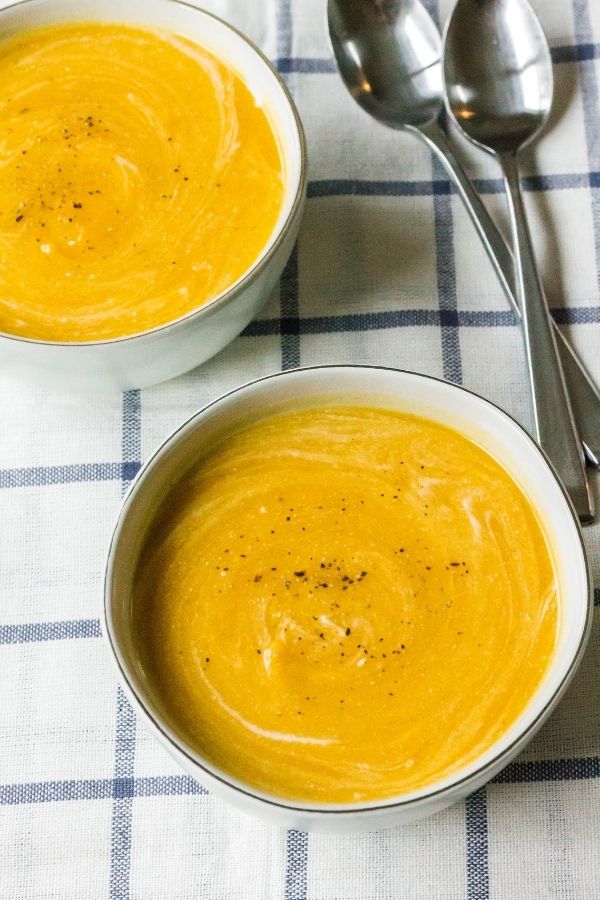 two white bowls filled with Instant Pot butternut squash soup, topped with cracked black pepper. Two spoons are nearby on the blue and white checked tablecloth.