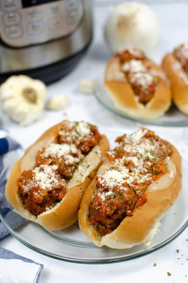 two clear plates, each with two pressure cooker meatball subs on it. Each sub has provolone cheese, meatballs, sauce, and grated parmesan cheese. An Instant Pot is in the background.