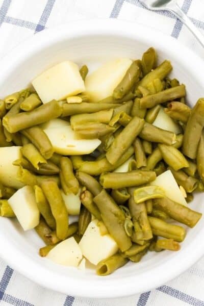 cooked canned green beans and potatoes are in a white bowl on a blue and white cloth, after being cooked in the pressure cooker