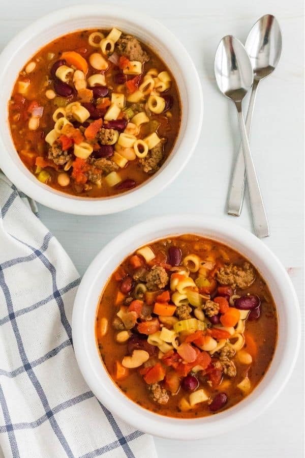 Two white bowls filled with Instant Pot pasta fagioli, next to two spoons and a white and blue checked dish cloth