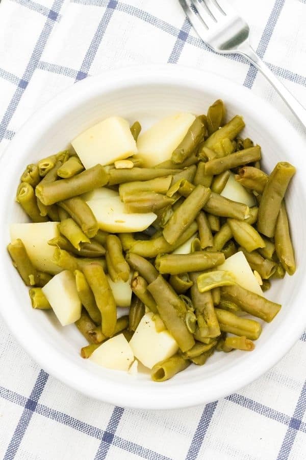 a white dish filled with Instapot green beans and potatoes, sitting on a blue and white checked surface, with a fork nearby