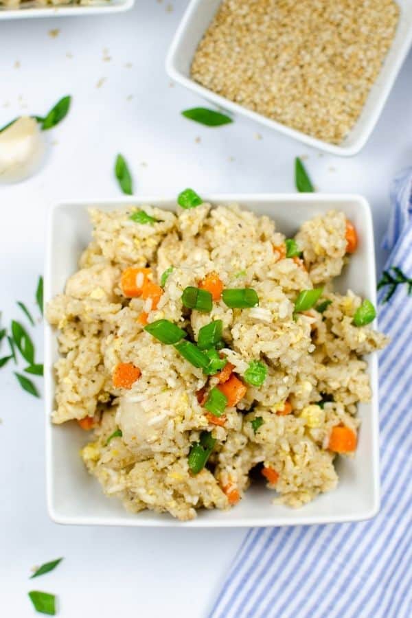 a white bowl of chicken fried rice cooked in the pressure cooker, next to a blue and white striped towel and a bowel of sesame seeds for garnish