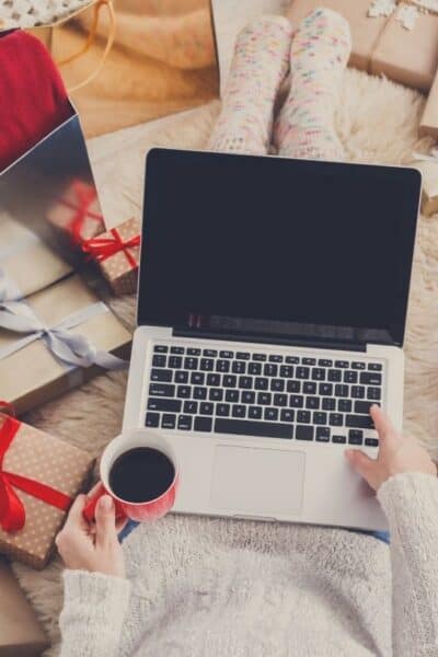 woman shopping on her laptop while holding a cup of coffee, with multiple Christmas presents surrounding her