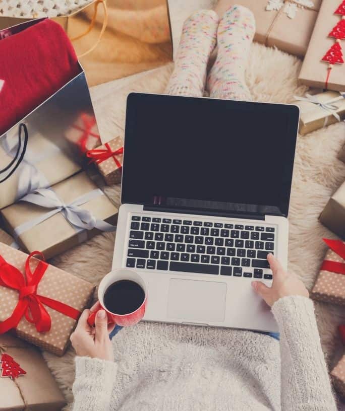 woman shopping on her laptop while holding a cup of coffee, with multiple Christmas presents surrounding her