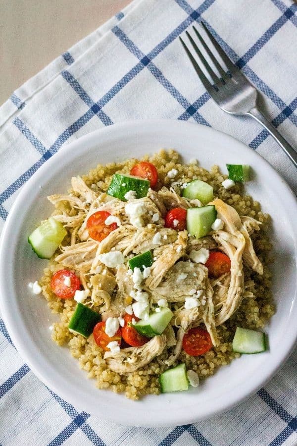 A white plate of Instant Pot Greek chicken served over quinoa and topped with cucumbers, tomatoes, and feta cheese, resting on a blue and white checked napkin, with a fork in the background.