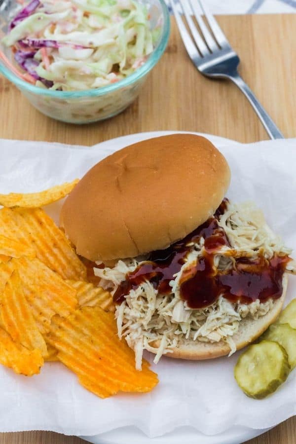 piece of parchment paper with a ranch chicken sandwich on it, as well as some potato chips, pickle slices, and a glass bowl of coleslaw.